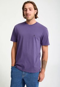 Lila bomulls-t-shirt med rund hals, korta ärmar och en liten logotyp på bröstet. Modellen bär den tillsammans med ljusblå jeans.