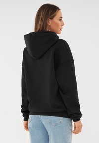 Sudadera de algodón negra con un corte relajado, gran capucha, puños y dobladillo de canalé. Se lleva con jeans de mezclilla azul claro.