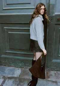 Mujer con cabello largo y ondulado, vestida con un suéter de punto gris, falda de cuero negra, botas negras, sosteniendo un bolso grande de ante marrón junto a una puerta verde.