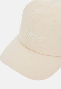 Casquette en coton beige avec une surface texturée, présentant un logo "GUESS" brodé à l'avant et une visière courbée.