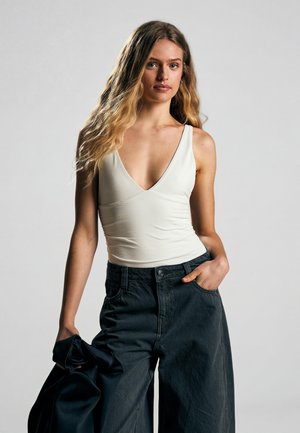 Mladá žena s dlhými vlnitými vlasmi má na sebe biele tielko s hlbokým výstrihom do V a voľné čierne nohavice, v jednej ruke drží čiernu bundu.