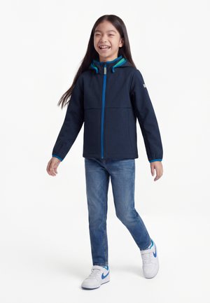 Niña joven caminando y sonriendo, vestida con una chaqueta con capucha azul marino, jeans azules y zapatillas blancas con detalles azules sobre un fondo blanco.