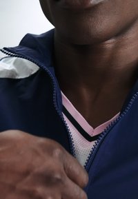 Primer plano de la mano de una persona cerrando la cremallera de una chaqueta azul marino sobre una camisa de rayas rosas y blancas, mostrando detalles del cuello y la parte inferior del rostro.