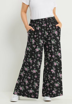 Kobieta ubrana w czarne szerokie spodnie z różowym kwiatowym wzorem i elastycznym paskiem, w połączeniu z białymi trampkami i białą bluzką z krótkim rękawem.