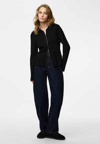 Cardigan nero con cerniera, colletto, corpetto aderente e maniche lunghe. Indossato con jeans blu scuro a gamba larga e scarpe nere slip-on.