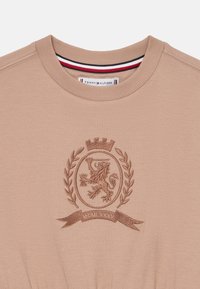 T-shirt in cotone beige con scollatura rotonda, emblema ricamato con un leone e alloro, e un banner con la scritta "MCMLXXXV". Il colletto interno presenta strisce rosse, bianche e blu.