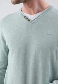 Pull vert clair en V avec trois boutons en bois et une finition texturée. Présente des coutures visibles et un logo discret sur la poitrine.