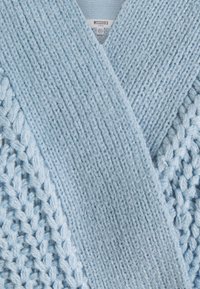 Cardigan bleu clair en tricot avec un motif texturé, présentant un large col en V et des motifs de points détaillés. Étiquettes visibles à l'intérieur.