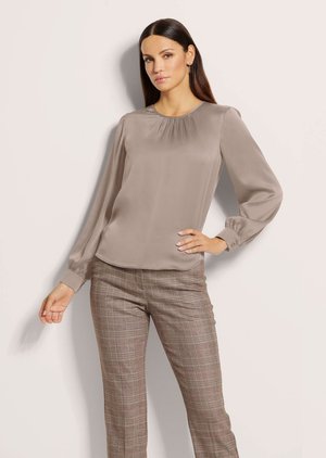 Femme aux longs cheveux foncés portant un chemisier taupe à manches longues et un pantalon marron à carreaux, posant avec une main sur la hanche sur un fond neutre.