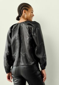 Juoda bluzonė su ilgomis pūkuotomis rankovėmis, satino tipo tekstūra, su sidabrinėmis juostelėmis siuvinėjimo detalėmis. Uždarymas gale su raukinių akcentu.