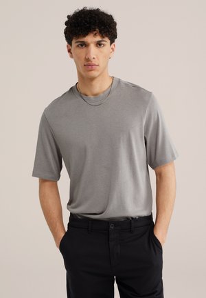 Giovane uomo con capelli neri ricci che indossa una maglietta grigia a maniche corte e pantaloni neri con le mani nelle tasche, in piedi davanti a uno sfondo uniforme.