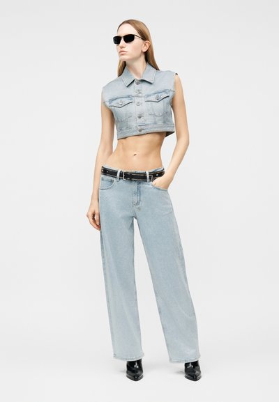 Γυναίκα με αμάνικο κοντό τζιν μπουφάν, τζιν παντελόνι με φαρδιά μπατζάκια και ζώνη, μαύρες μυτερές μπότες και γυαλιά ηλίου, στέκεται με το χέρι στην τσέπη.