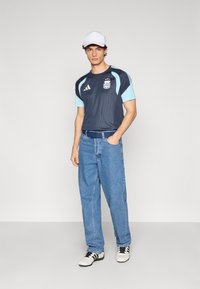 Navy mesh athletic shirt with teal accents, light blue sleeve panels, blue jeans, and black-and-white athletic shoes. White cap.