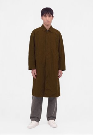 Jeune homme debout face à l'avant portant un long manteau marron, un pantalon gris ample et des baskets blanches sur un fond blanc uni.