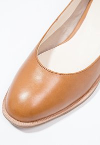 Gros plan d'une chaussure plate en cuir tanné avec un bout carré arrondi et une semelle intérieure beige rembourrée sur un fond blanc.