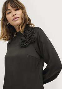 Zwarte blouse met lange mouwen, hoge hals en driedimensionale stoffen rozen op de linkerschouder. Gladde textuur en getailleerd ontwerp.