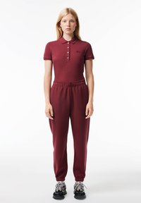 Frau in einem passenden burgunderroten Polo-Shirt und Jogginghose mit schwarzen und weißen Sneakern, die vor einem weißen Hintergrund steht.