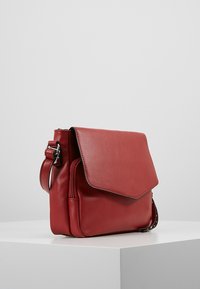 Červená kožená crossbody taška s předním klopovým víkem a zipovou kapsou, nastavitelné ramenní popruhy se stříbrnou sponou, vystavená na bílé podstavě na šedém pozadí.