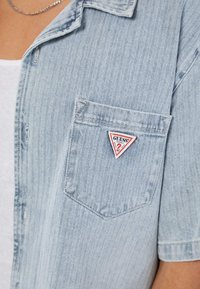 Lichtblauw denim overhemd met korte mouwen en een voorzak, voorzien van een rood en wit driehoekig Guess merklogo-patch.