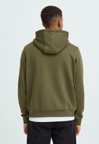 Olive green hoodie with a relaxed fit, adjustable drawstring hood, ribbed cuffs, and hem. Worn over a white shirt and black pants.
