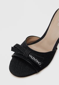Zapato de tacón alto de denim negro con un diseño de punta abierta y suave, que presenta un lazo de tela y detalle de logo en el lateral. Forma estructurada con líneas elegantes.