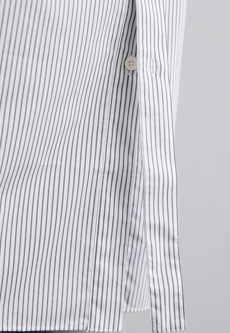 Chemise blanche avec de fines rayures verticales grises, dotée d'une fermeture à boutons et d'un ourlet arrondi. Le tissu paraît léger et lisse.