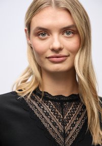 Junge Frau mit blonden Haaren, die ein schwarzes Oberteil mit einem detaillierten, spitzen Spitzenkragen trägt, schaut direkt in die Kamera.