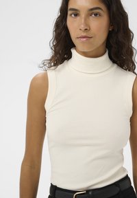 Jonge vrouw met krullend haar, gekleed in een mouwloos wit geribd coltruitje en een zwarte riem, tegen een effen achtergrond.