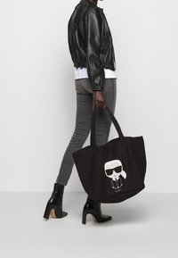 Osoba oblečená v černé kožené bundě, šedých slim kalhotách, černých botách na podpatku, nese velkou černou tašku s potiskem kreslené postavy.