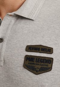 Camisa polo gris con cuello corto, que presenta dos parches bordados: "FLYING RELIC" encima de "PME LEGEND" con un gráfico de alas. Texto en colores oscuros.