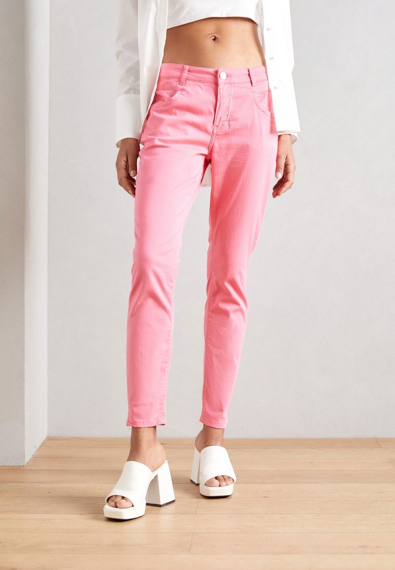 Jean rose ajusté en mélange coton-spandex, doté de poches, d'une fermeture à bouton et d'une texture lisse. Porté avec des sandales blanches à talons blocs.