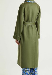 Manteau long en laine vert olive avec une taille ceinturée et des manches larges. Il est doté d'une doublure intérieure à motif floral et d'une coupe droite.