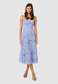 Abito midi azzurro chiaro con motivi tagliati al laser, spalline regolabili e una silhouette fluida a forma di A. Sandali completano l'outfit.