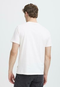 Camiseta de algodón de manga corta y cuello redondo en color blanco roto. Presenta un ajuste relajado, una textura suave y una etiqueta en el lado inferior izquierdo. Se muestra la vista trasera.