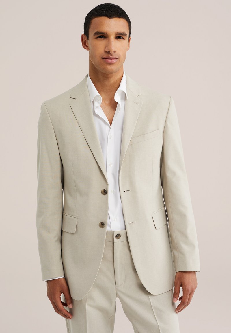 Homme portant un costume beige avec une chemise blanche, debout devant un fond clair uni, face à l'avant avec une expression neutre.
