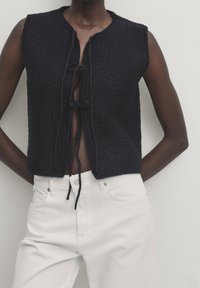 Gilet lavorato a maglia blu navy con una finitura testurizzata, maniche corte e chiusura anteriore con fiocco, abbinato a pantaloni bianchi.