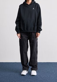 Sudadera negra de algodón con capucha, bolsillo canguro delantero y logo blanco. Vaqueros oscuros de corte holgado con detalles dorados. Zapatillas blancas.