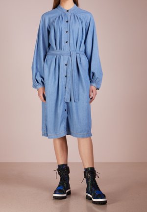 Hellblaues Denimhemdkleid mit hohem Kragen, Knopfleiste und langen Ärmeln. Mit gebundenem Taillenband und elastischen Bündchen. Kombiniert mit schwarzen Kampfstiefeln.