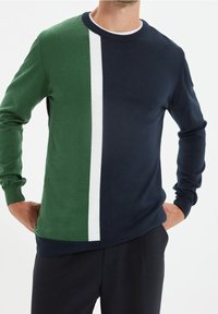 Pull-over en tricot avec un panneau gauche vert et un panneau droit marine, accentué d'une bande verticale blanche. Fabriqué en tissu tricoté avec des manches longues.