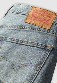 Calças de ganga azul claro com um remendo de couro rotulado "Levi Strauss & Co.", com texto vermelho e dois cavalos. Inclui bolsos traseiros padrão.