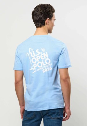 Man draagt een lichtblauw t-shirt met witte tekst "U.S. Open Polo Championship 2026" en een polo-knuppel afbeelding op de rug.