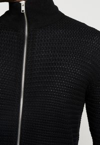 Svart strukturerad stickad tröja med hög krage och silverdragkedja som går ner mitt fram.