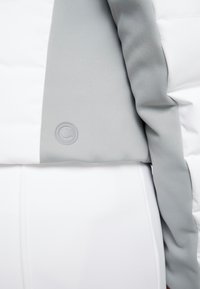 Vadderad jacka i vitt med grå accenter. Inkluderar en strukturerad grå panel och en cirkulär logotyp på ärmen.