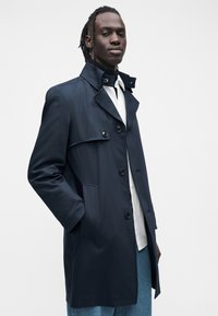 Palton bleumarin cu guler, cu două rânduri de nasturi în față și buzunare laterale. Țesătura pare netedă, cu croială structurată și design personalizat.