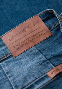 Tissu en denim avec une étiquette en cuir marron portant un texte embossé. L'étiquette indique "Ombre" et comprend un slogan sur le style. Couture contrastée visible.