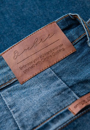 Tissu en denim avec une étiquette en cuir marron portant un texte embossé. L'étiquette indique "Ombre" et comprend un slogan sur le style. Couture contrastée visible.