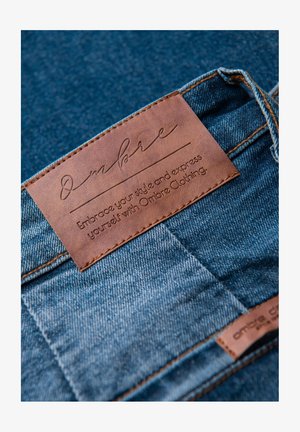 Tessuto di denim con un'etichetta in pelle marrone caratterizzata da testo in rilievo. L'etichetta riporta la scritta "Ombre" e include uno slogan sullo stile. Cuciture a contrasto visibili.
