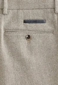 Lysebrunt tekstureret stof af bukser med en baglomme med brun knaplukning og en sort detalje.