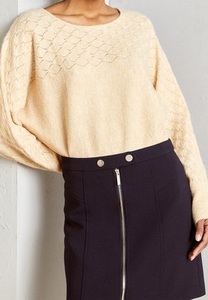 Femme portant un pull en maille beige à motif losange et une jupe noire avec une fermeture éclair à l'avant et deux boutons argentés à la taille.