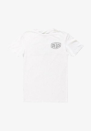Camiseta de algodón blanca con mangas cortas y cuello redondo. Presenta un logo de escudo gris con "DEUS" en el pecho izquierdo. Diseño simple.